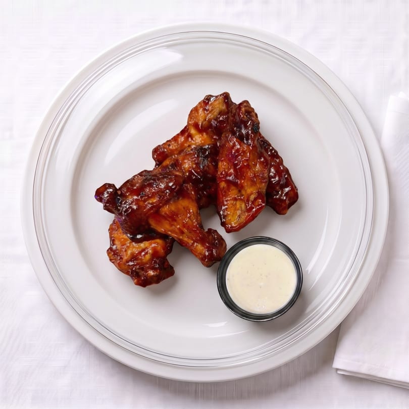 Glazed chicken wings with dipping sauce on a white plate, next to a white napkin.
