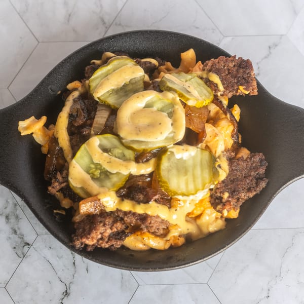 Loaded fries topped with crispy ground beef, sliced pickles, grilled onions, and creamy cheese sauce in a black skillet.