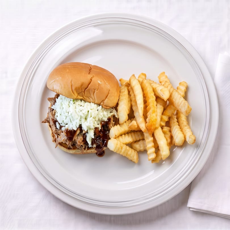 Pulled pork sandwich topped with coleslaw and BBQ sauce, served with crinkle-cut fries on a white plate.