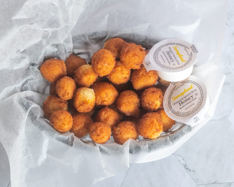 Crispy golden-brown hushpuppies in a paper-lined basket with two Sunnyland whipped honey cups.