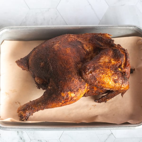 Whole roasted chicken with a dark seasoned crust resting on parchment in a metal baking tray.