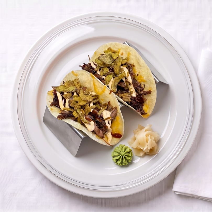 Two tacos filled with shredded meat, crispy toppings, and sauce, served with wasabi and pickled ginger.