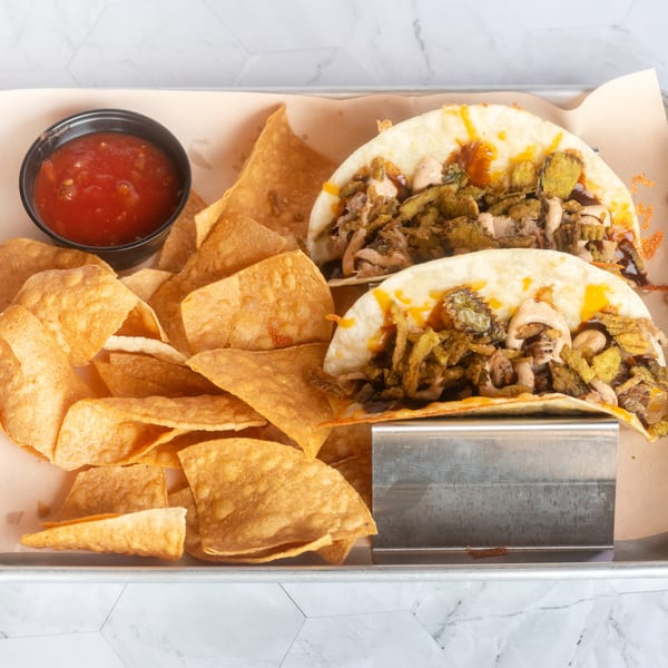 Two cheesy tacos topped with shredded meat and fried vegetables, served with tortilla chips and red salsa on a metal tray.