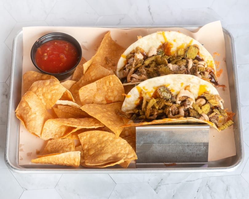 Two tacos with crispy toppings, tortilla chips, and salsa on a metal tray.