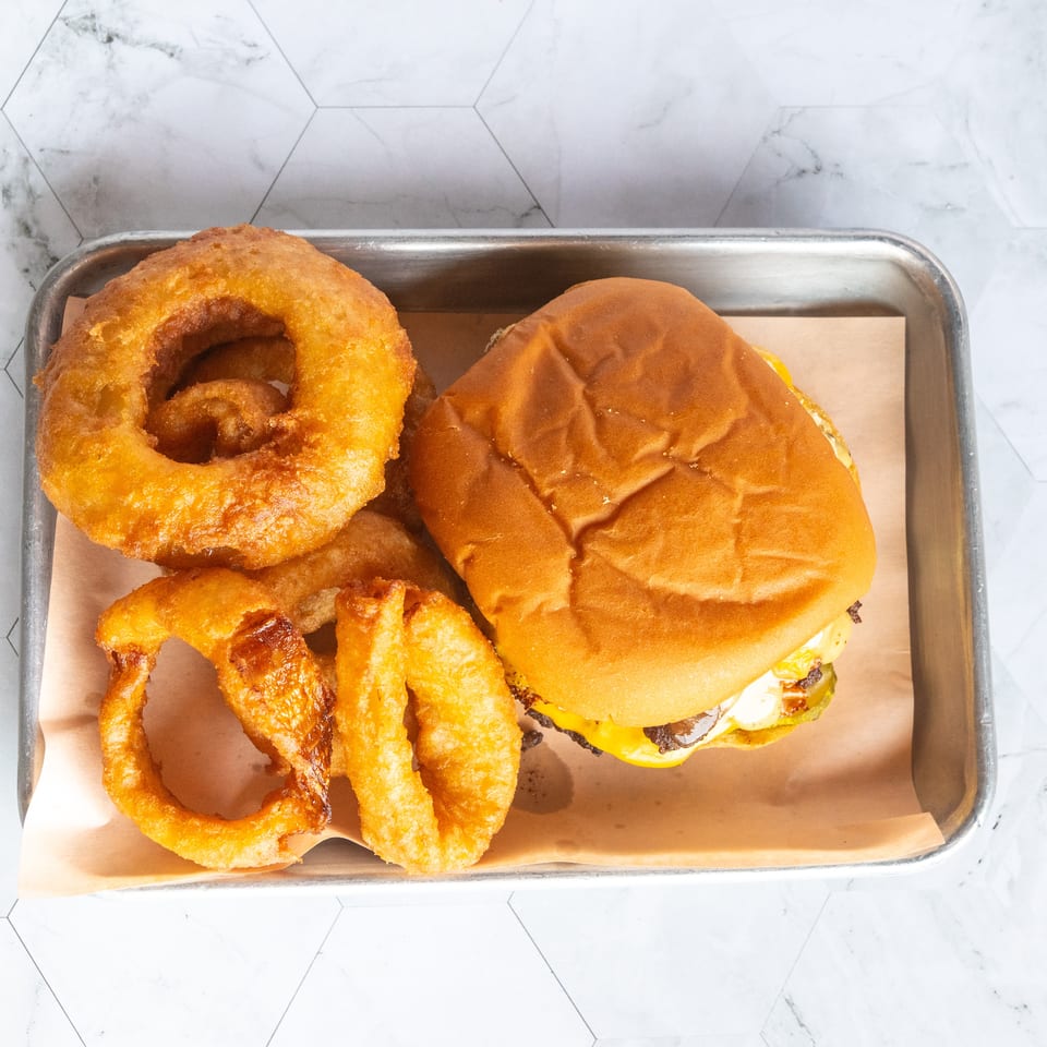 Burger with melted cheese, crispy fries, and golden onion rings served on a parchment-lined metal tray.