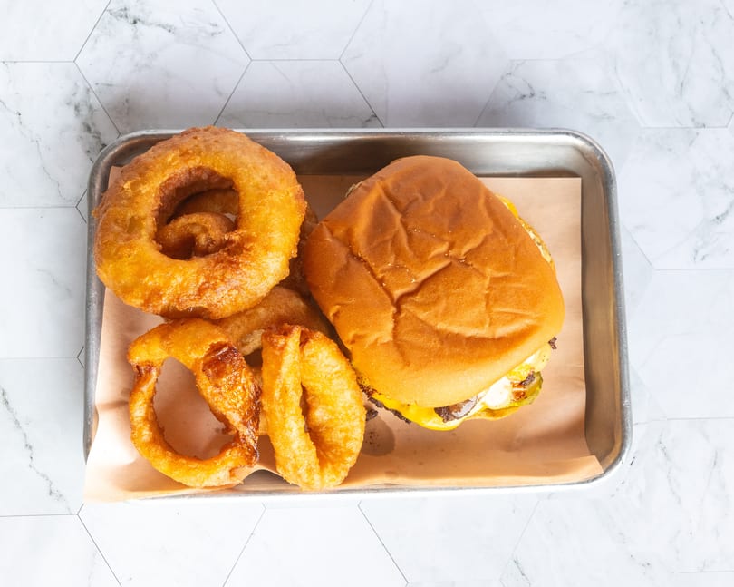 Golden onion rings and a cheeseburger with pickles on a metal tray.