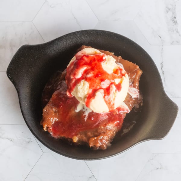 Warm dessert topped with vanilla ice cream and red berry sauce, served in a black skillet on a marble surface.