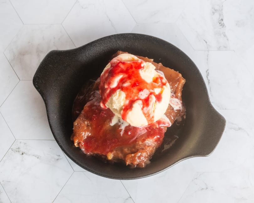 Warm fruit cobbler with melting vanilla ice cream and strawberry sauce in a dark bowl.