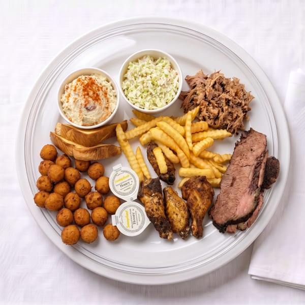 A large platter filled with BBQ pulled pork, brisket, chicken wings, fries, hush puppies, coleslaw, and potato salad.