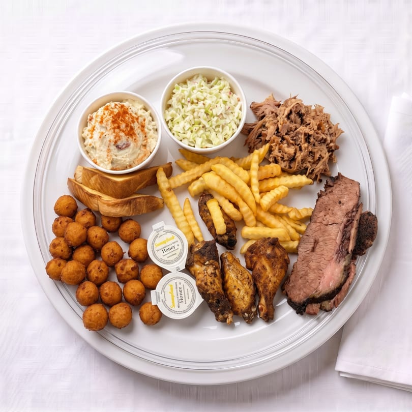 A large platter filled with BBQ pulled pork, brisket, chicken wings, fries, hush puppies, coleslaw, and potato salad.