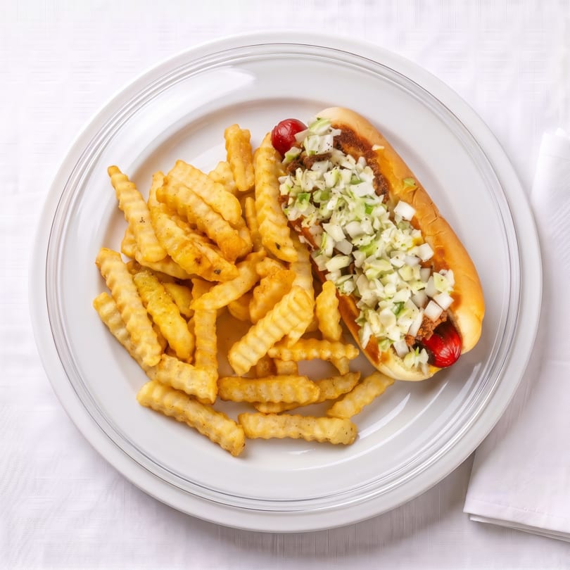 A hot dog with chili, coleslaw, and a red topping, served with crinkle-cut fries on a white plate.