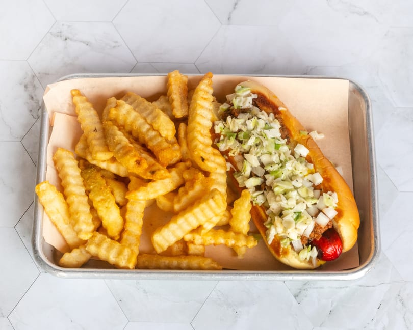 Chili dog topped with coleslaw and onions, served with crinkle-cut fries on a metal tray.